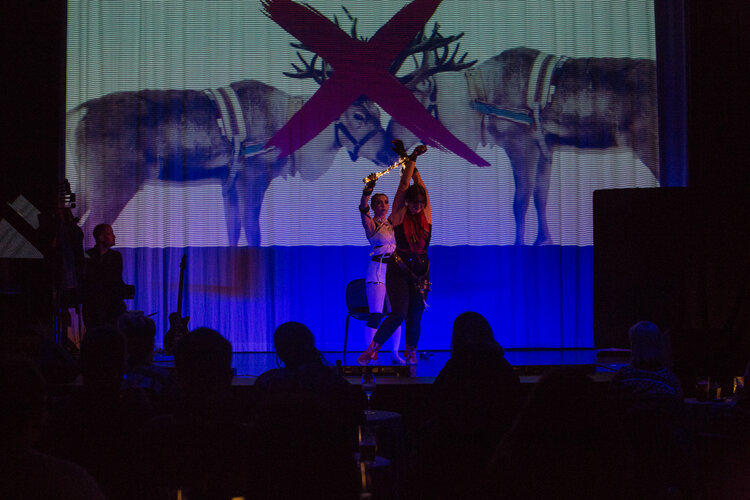 To utøvere på en scene under en forestilling. Lerret med bilde av reinsdyr i bakgrunnen.