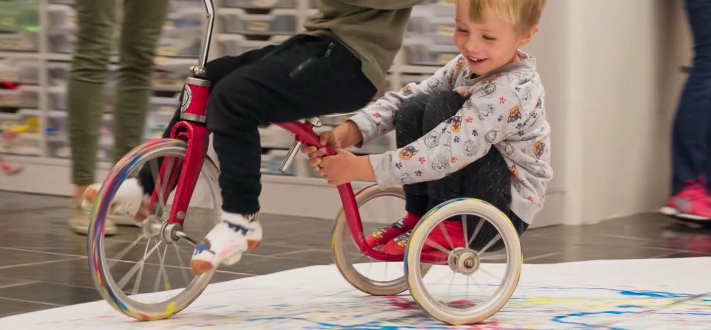 Gutt sitter bakpå en som sykler inne. De sykler rundt på et stort papir, maler med sykkelhjulene.