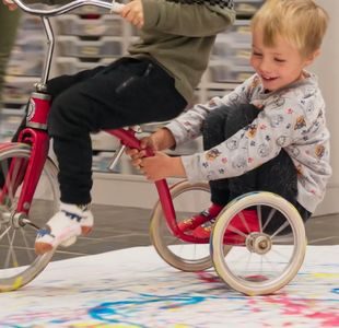 Gutt sitter bakpå en som sykler inne. De sykler rundt på et stort papir, maler med sykkelhjulene.
