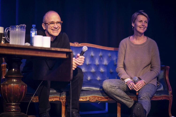 David Chocron og Gine Heien smiler, sitter i en sofa på scena. Chocron med mikrofon i hånda.