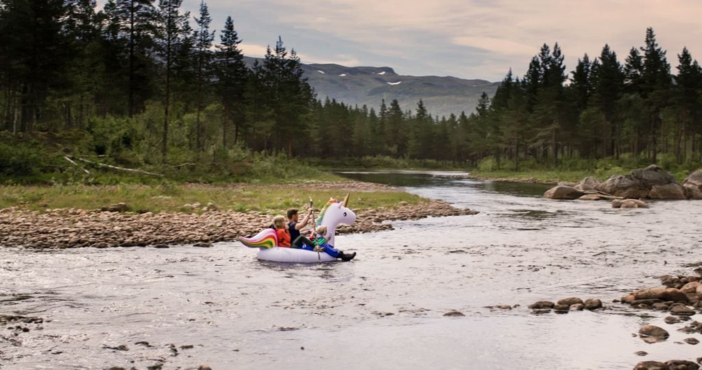 En far og to barn sitter i en stor, oppblåsbar enhjørning som flyter på en elv