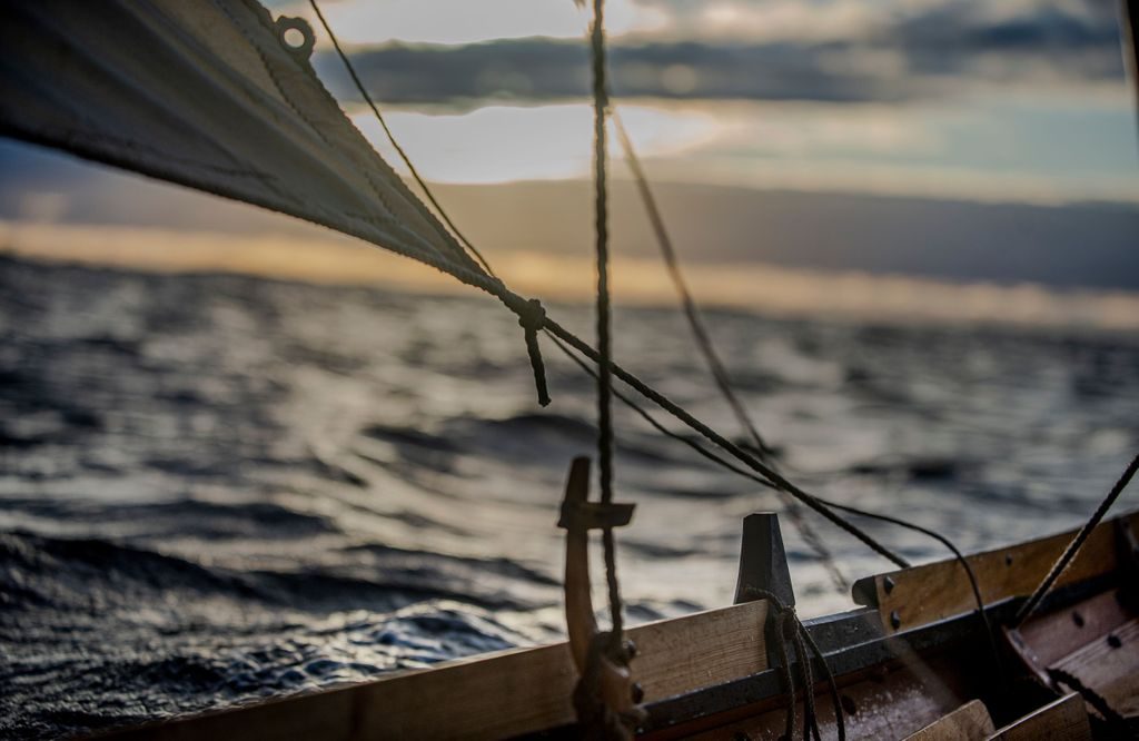 Vi ser deler av en trebåt med seil foran et sjølandskap i solnedgang
