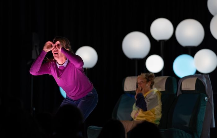 To kvinner på en scene. En kvinne i lilla genser holder fingrene opp som briller foran øynene.