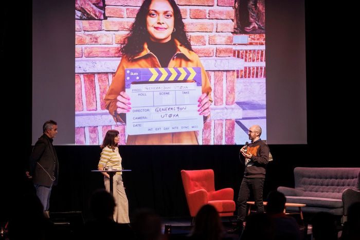 En programleder og to personer står på en scene og presenterer en film for publikum