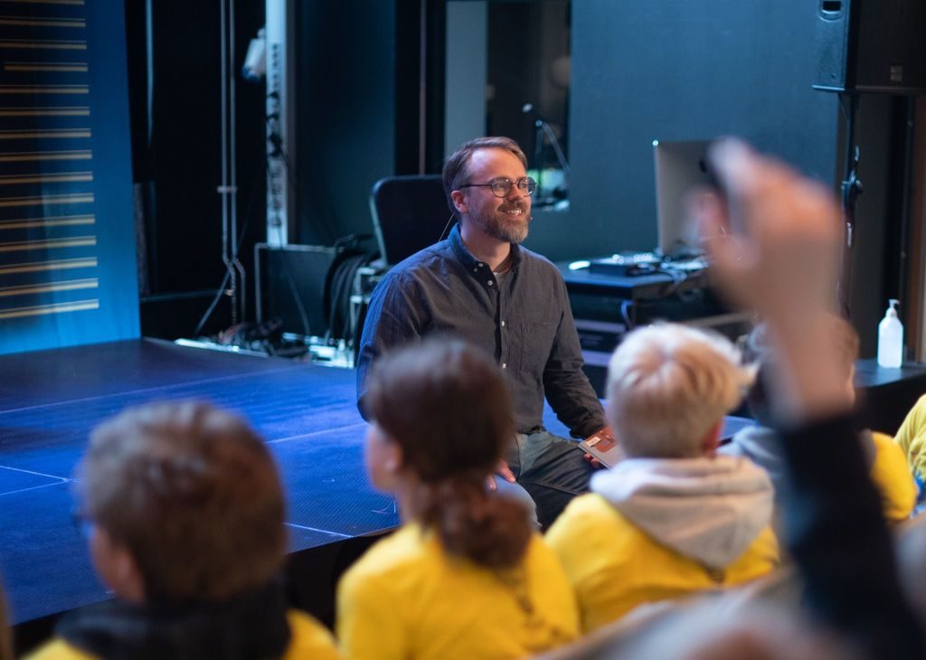 En mann sitter på kanten av en scene og smiler mot sitt publikum, som er flere barn med gule t-skjorter og ryggen mot kamera. En av dem har hånden i været.
