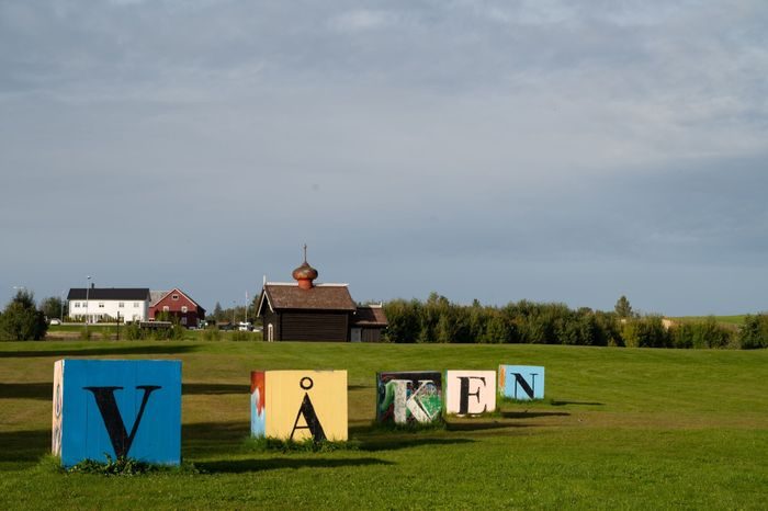 Treblokker med bokstaver som utgjør ordet "våken" står på en gressplen foran en gård