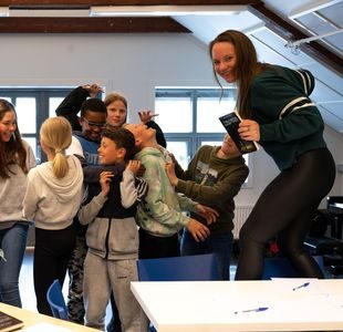 Forfatter Mari Moen Holsve står på en stol foran en gruppe elever som gjør ulike bevegelser og smiler