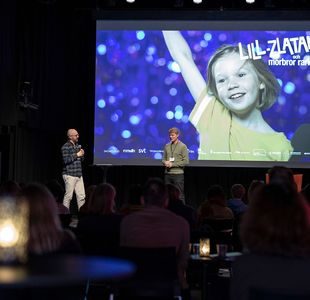 To menn snakker på en scene foran et mørklagt publikum. Bak dem er det en storskjerm med et bilde av en smilende jente som strekker en arm i været.