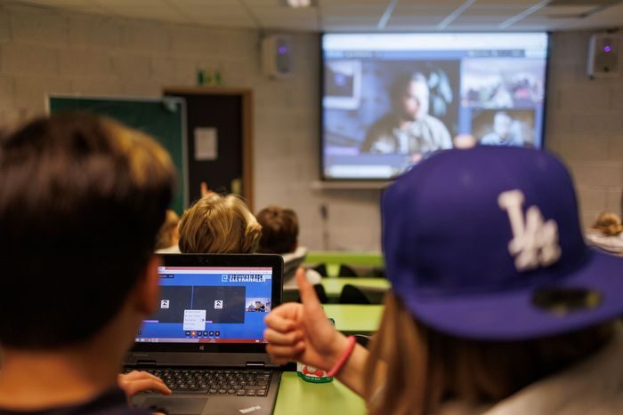 To elever sitter med laptop, en viser tommelen opp, skjerm med utøvere i bakgrunnen.