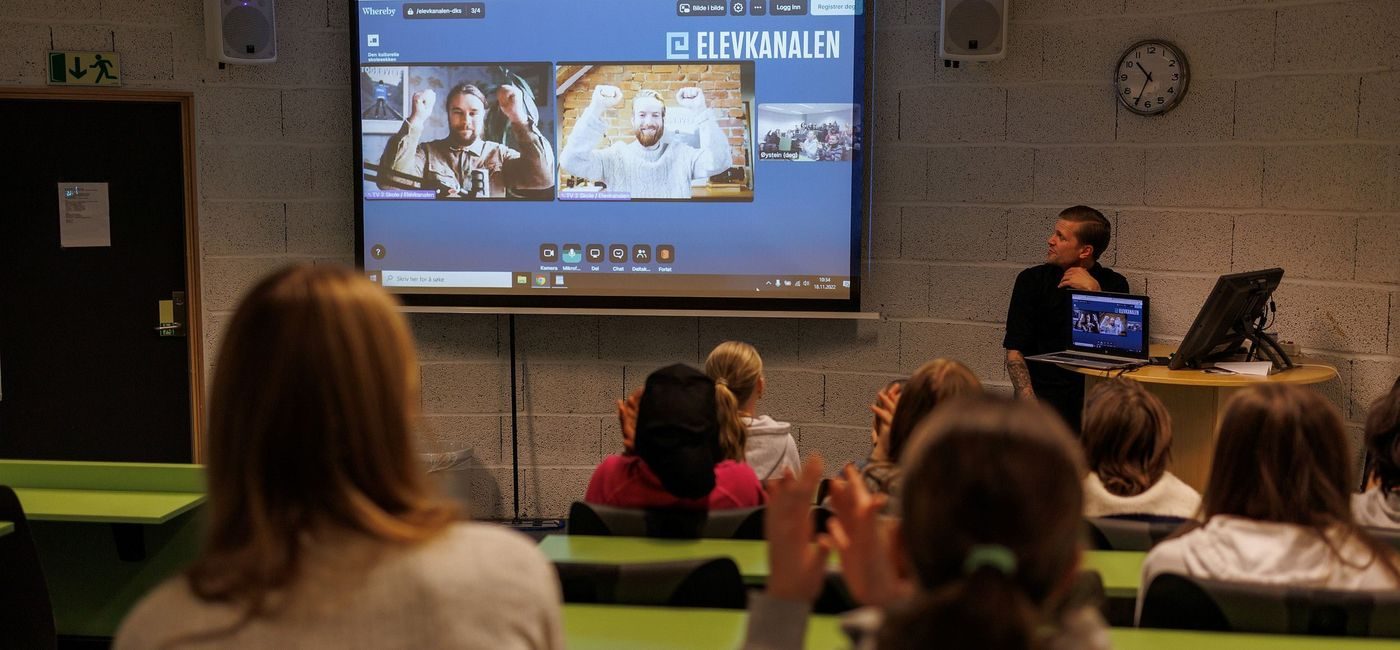 To utøvere i videovinduer på skjerm strekker hendene i været, elever i auditoriet klapper.