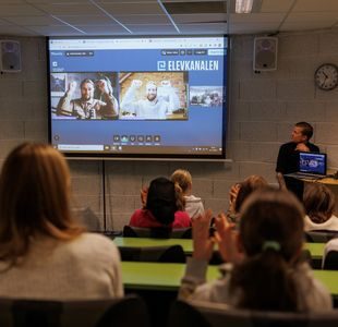 To utøvere i videovinduer på skjerm strekker hendene i været, elever i auditoriet klapper.