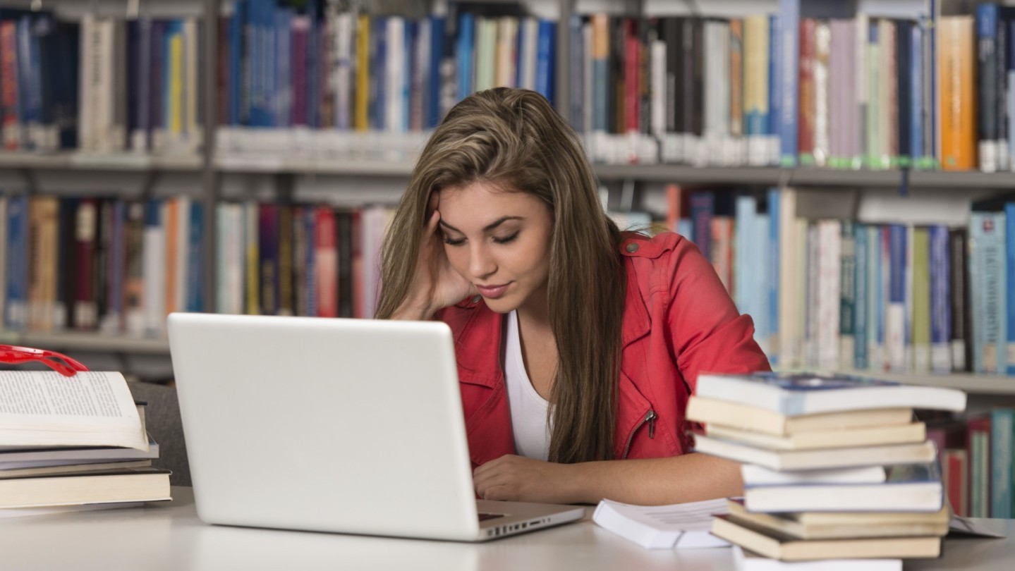Ung kvinnelig student jobber på datamaskin på biblioteket.