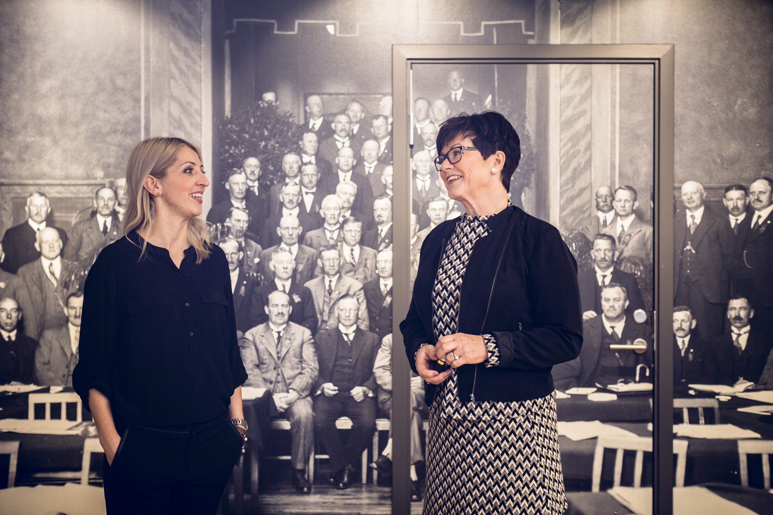 Jana Jarosova og Liv Spjeld By står sammen foran Ledernes vegg, dekorert med Ledernes første styre fra 1910.