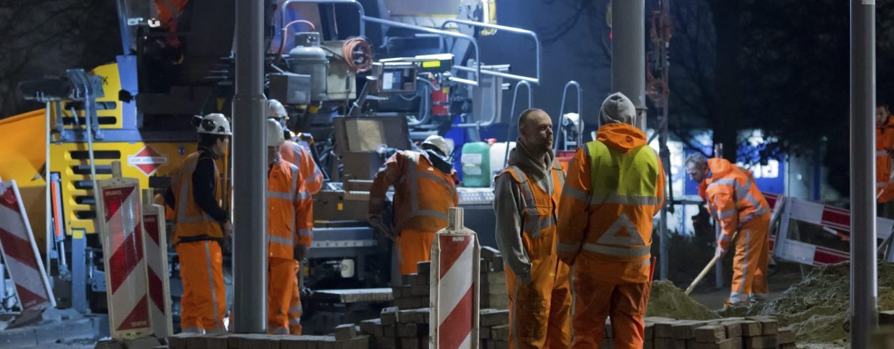 En gruppe anleggsarbeidere i orange klær jobber i mørket. En maskin og noen skilt er også med i bildet.