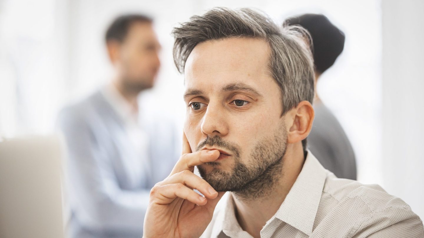 Mann på cirka 40 år, med grått hår og skjegg, sitter med en hånd foran munnen og ser tankefull ut. To andre anes i bakgrunnen.