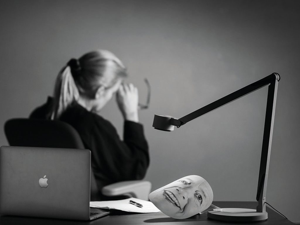 Balanse: Kvinne sitter vendt bort fra skrivebordet sitt og det ligger en maske på bordet med hennes smilende fjes på.
