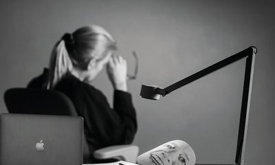 Balanse: Kvinne sitter vendt bort fra skrivebordet sitt og det ligger en maske på bordet med hennes smilende fjes på.
