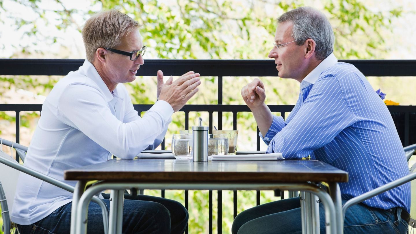 Sparre: To menn sitter og snakker ved et bord utendørs.