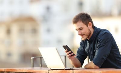 En mann står utendørs og lener seg til men mur, ser på mobilen og har laptopen foran seg. Illustrasjon til sak om arbeid og ferie i permittering
