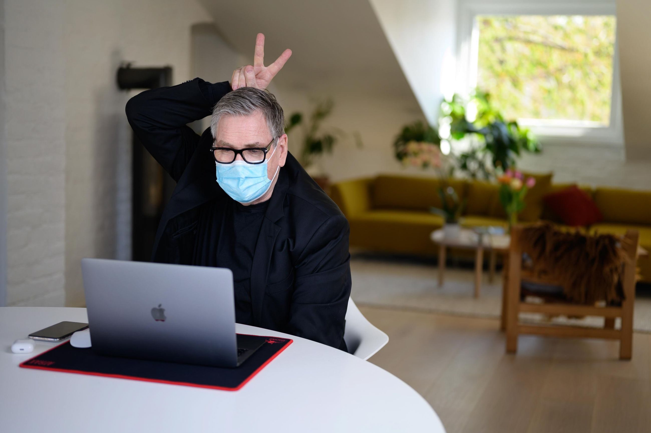 Mann foran laptop med munnbind som holder to fingre, i peacetegn, over eget hode