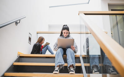 To jenter sitter i en trapp på en skole og ser på hver sin laptop. Bilde.
