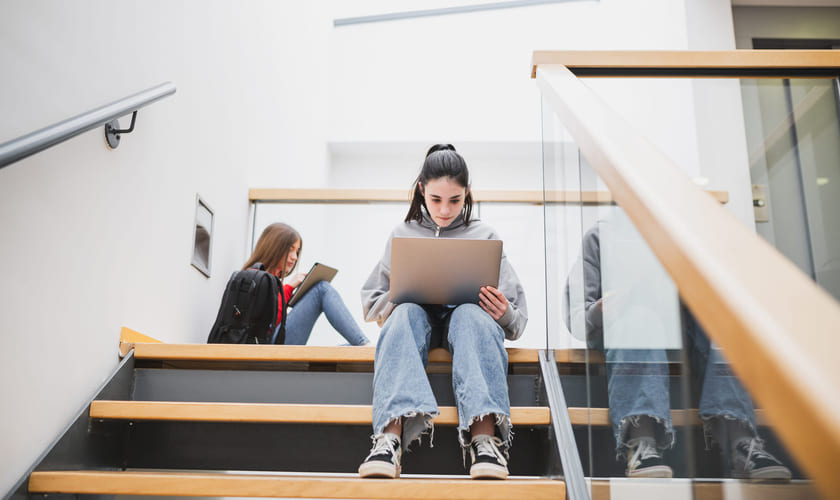 To jenter sitter i en trapp på en skole og ser på hver sin laptop. Bilde.