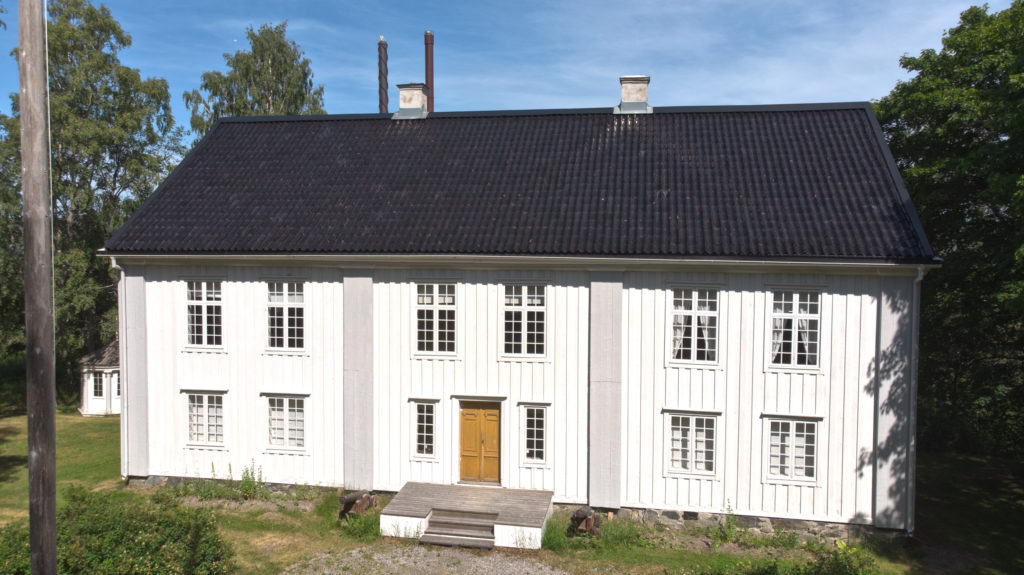 Ett vitt hus med svart tegeltak. På fasaden tolv fönster med spröjs. Ett par gula pardörrar. 