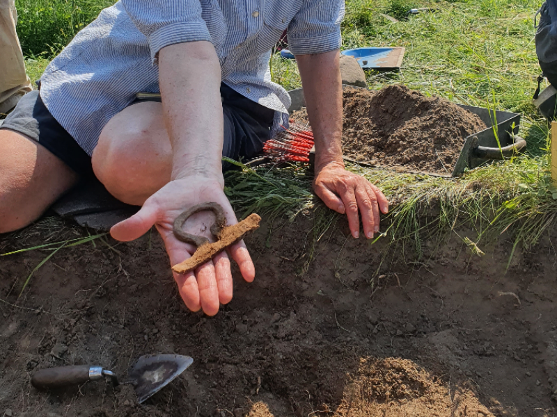 En person sträcker fram en hand med ett järnföremål i från en arkeologisk utgrävning